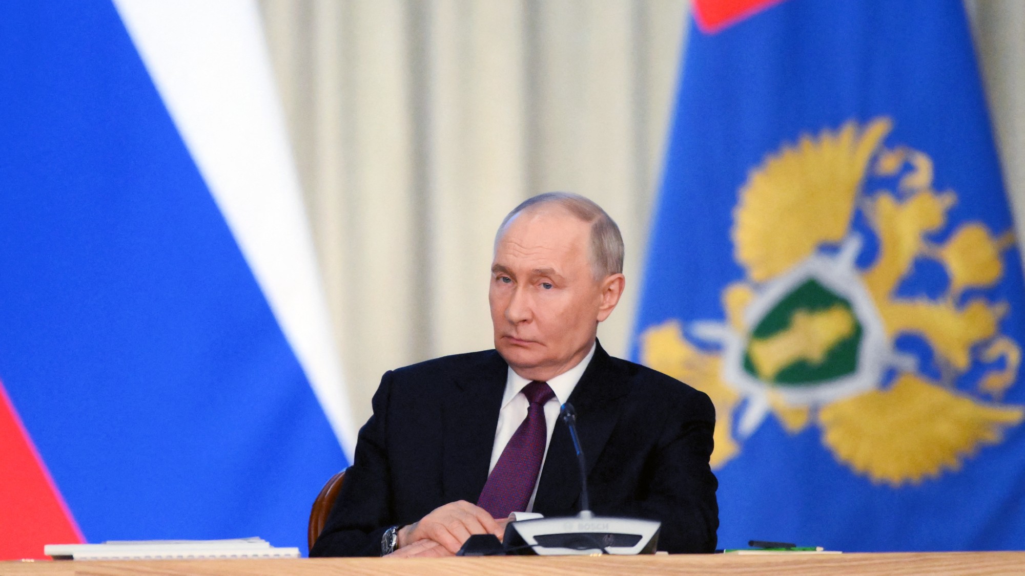 Vladimir Putin sitting at a table in front of a Russian flag