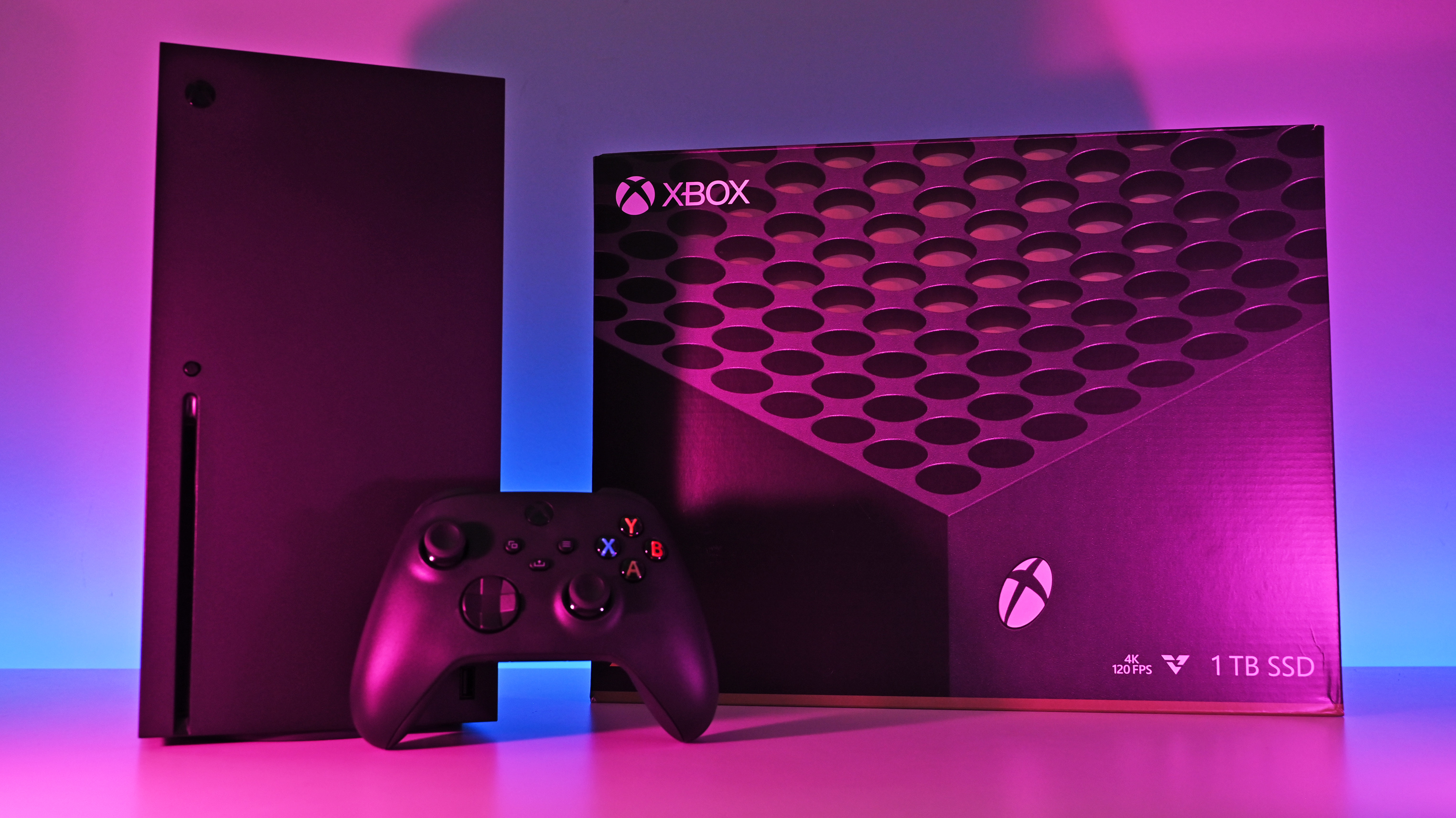 An Xbox Series X console, controller, and retail box standing under vibrant pink, purple, and blue atmospheric lighting.