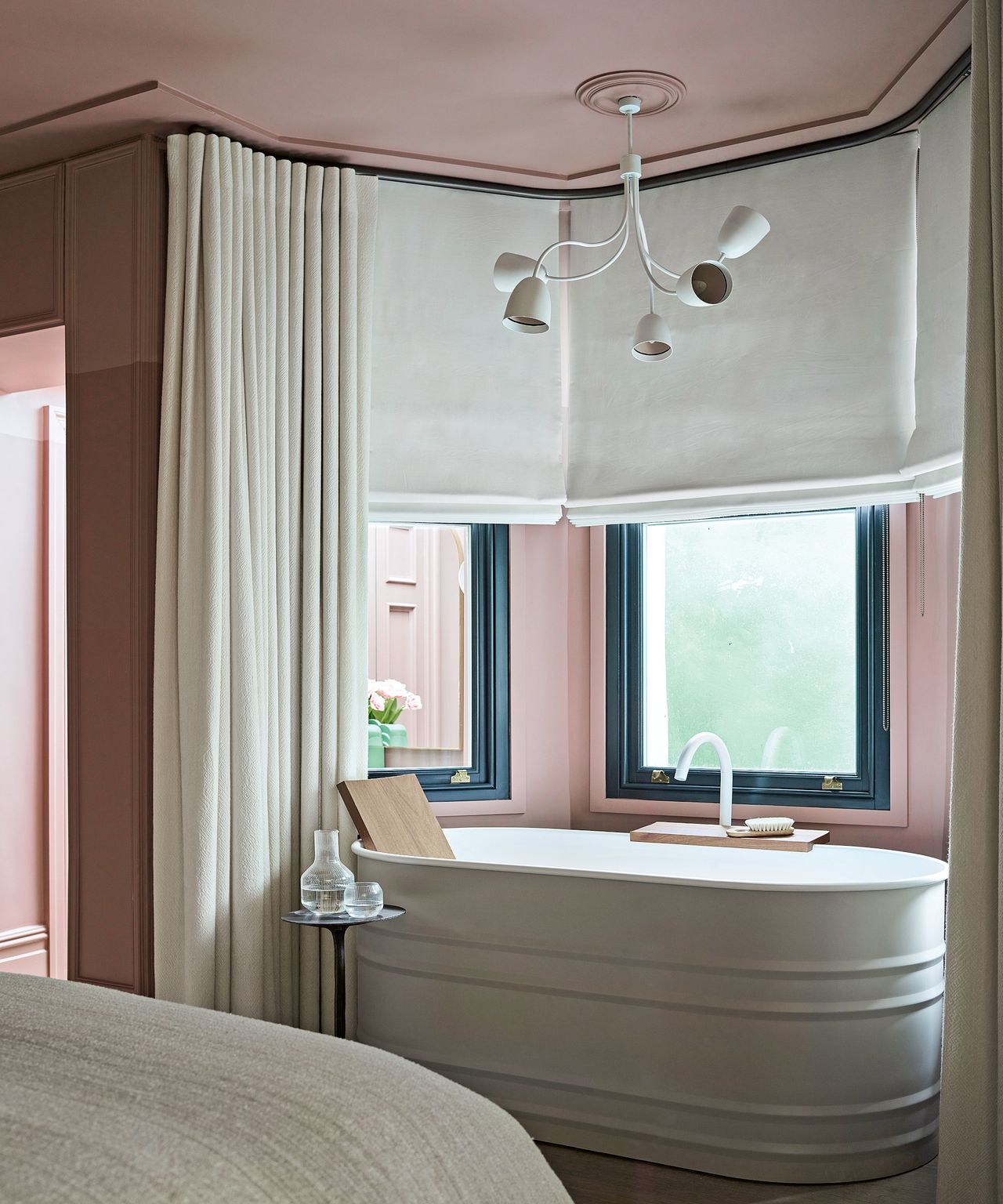 A bedroom with pink walls, a curved free-standing bathroom, and neutral drapes and drapes