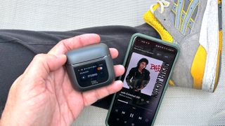 JBL Tour Pro 2 charging case in hand with a smartphones displaying MJ Bad album cover