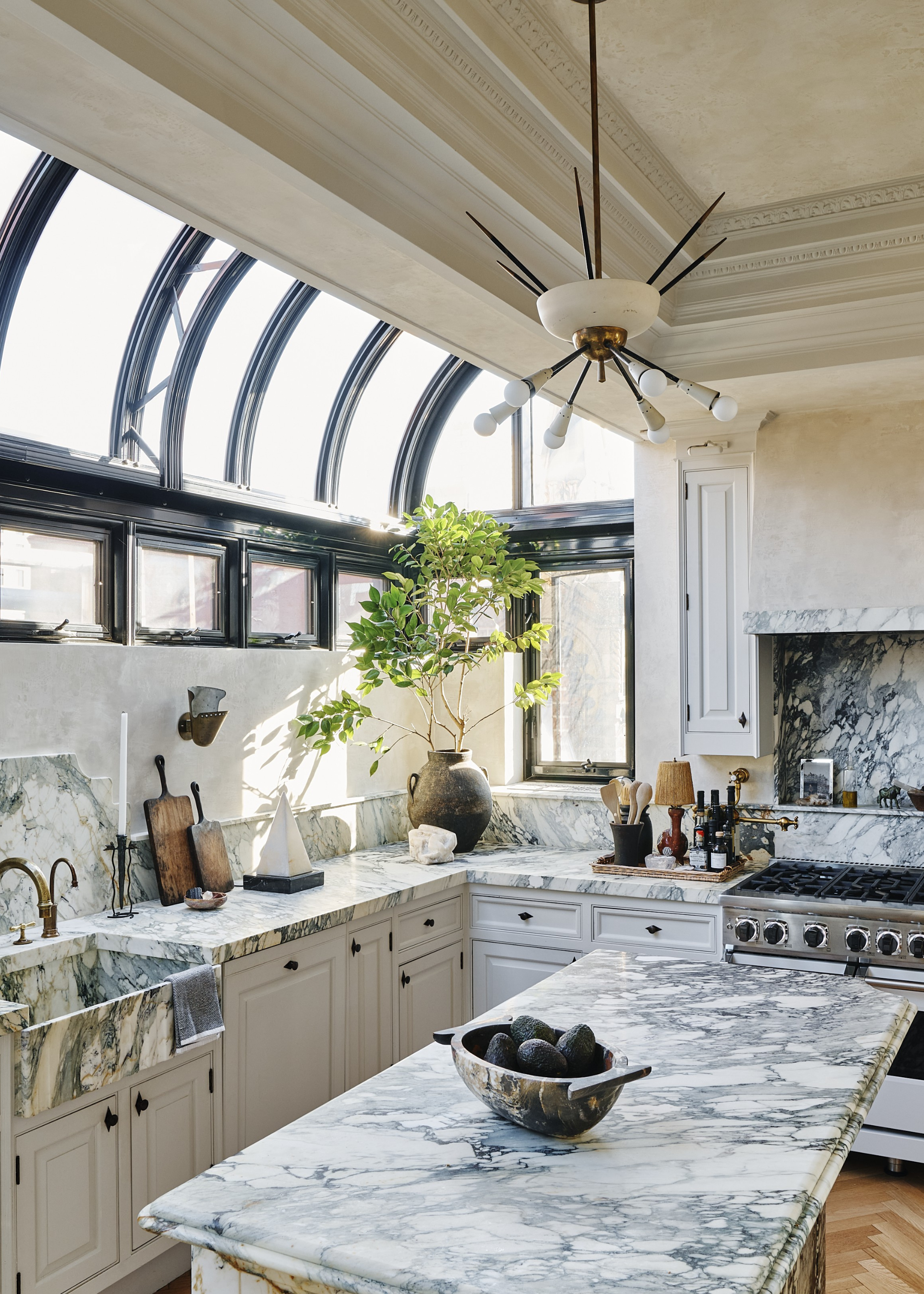 A contemporary kitchen with marble countertops and kitchen island with a pendant light above and glass-covered extension to expand the space with arched black-framed windows and a large plant sits on the countertop in the corner of the room