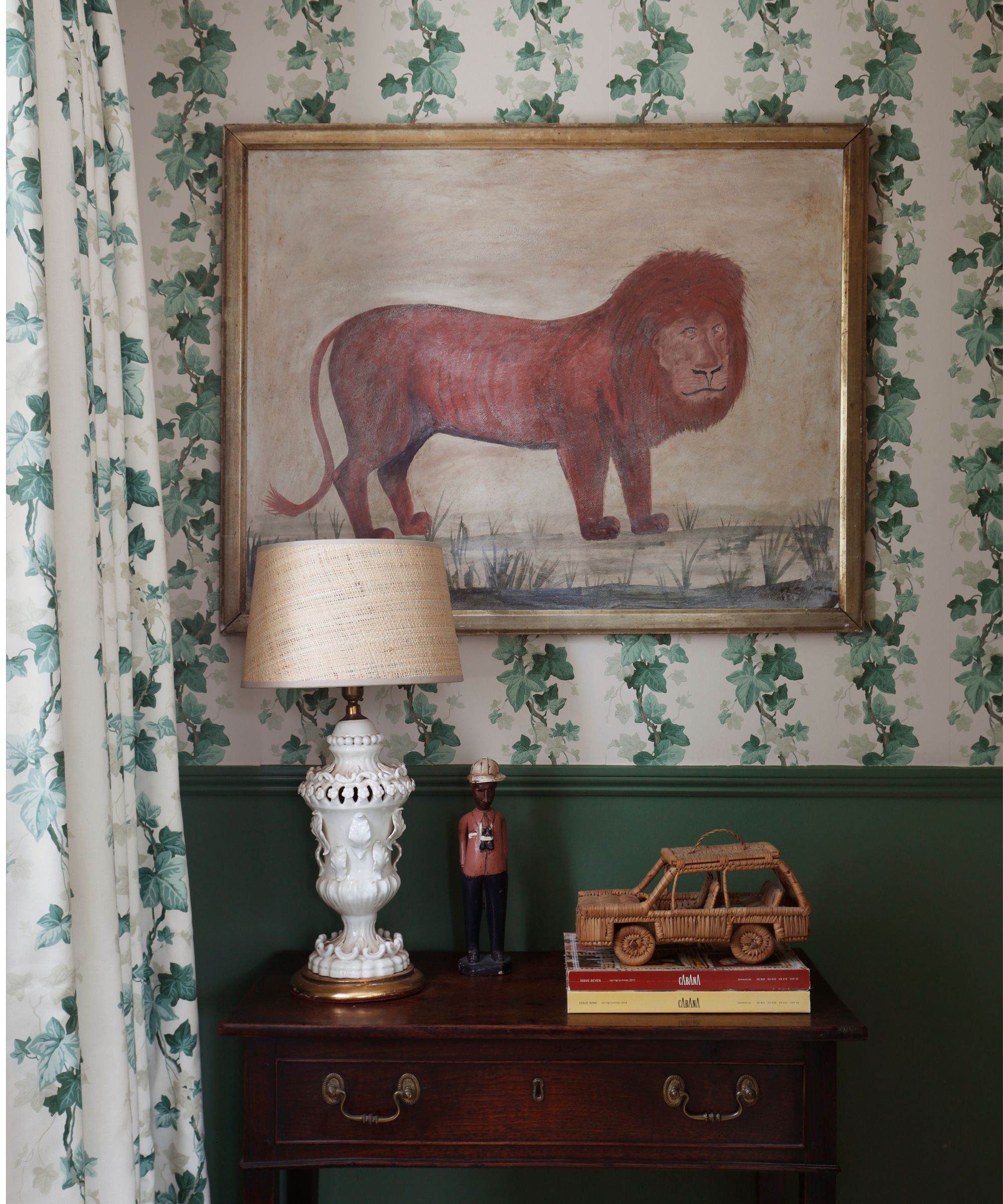 Hallway with ivy wallpaper and matching drapes, a Calke Green painted dado rail, a mahogany side chest with a white lamp and a toy car, and a large painting above it of a lion