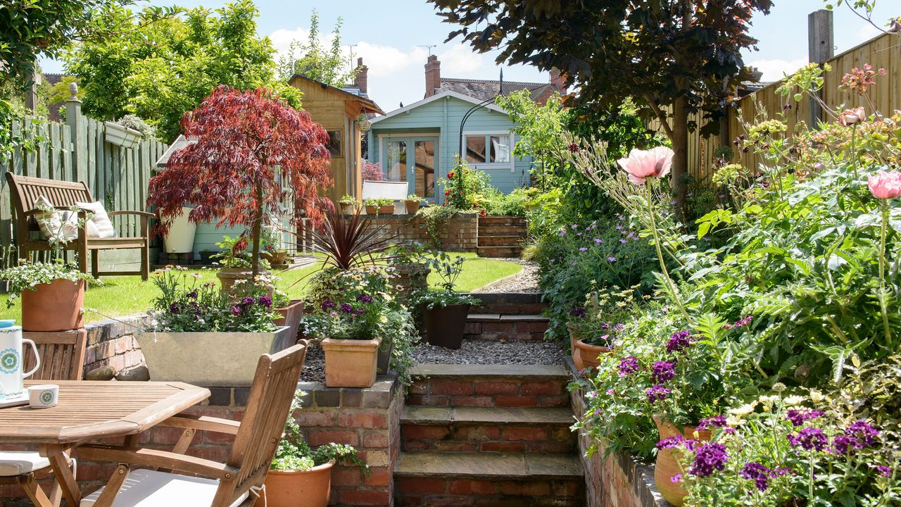 Garden in bloom with flowers, plants, steps and wooden table and chairs with blue shed in background