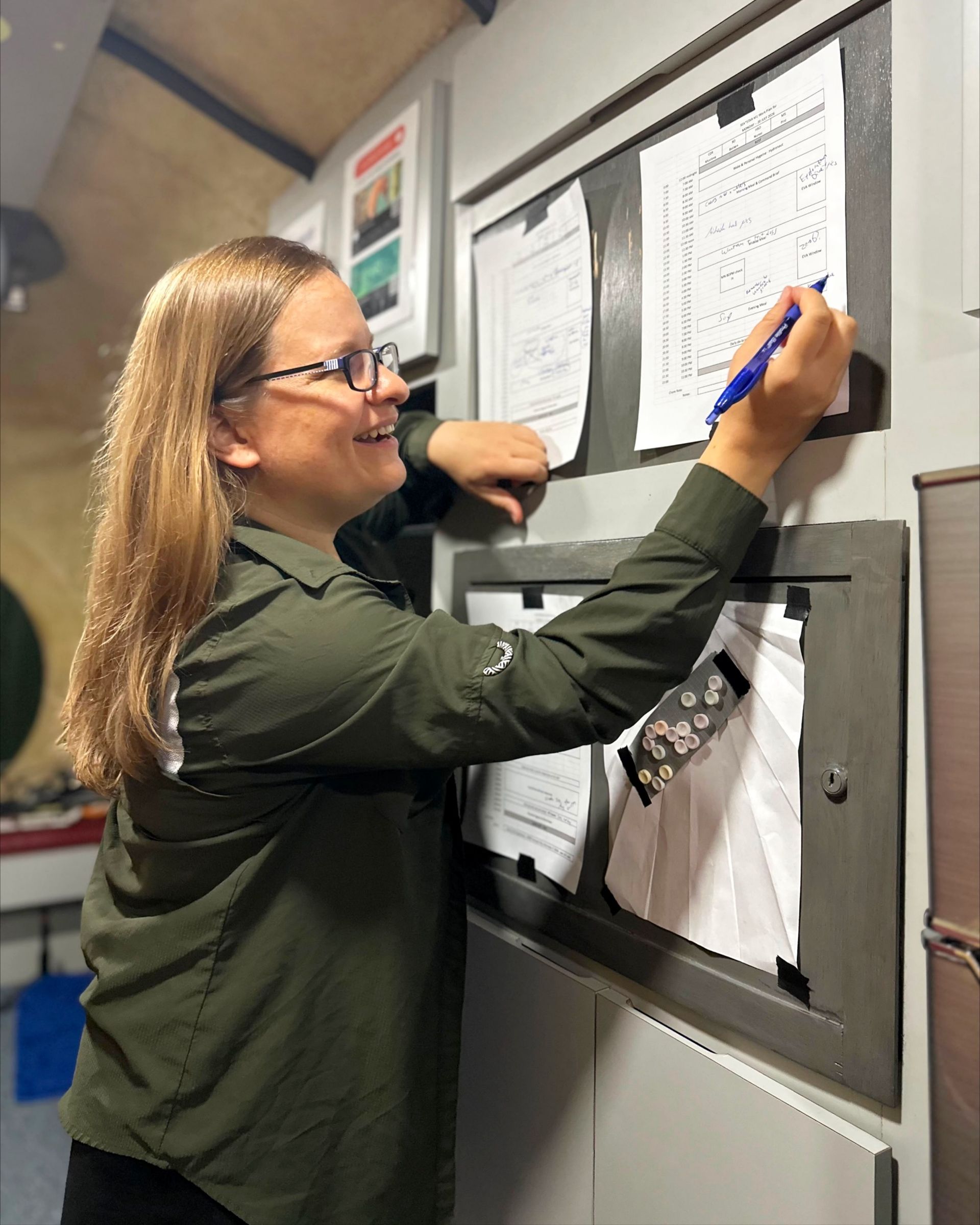 a smiling woman holding a blue pen works looks at a piece of paper pinned to the wall