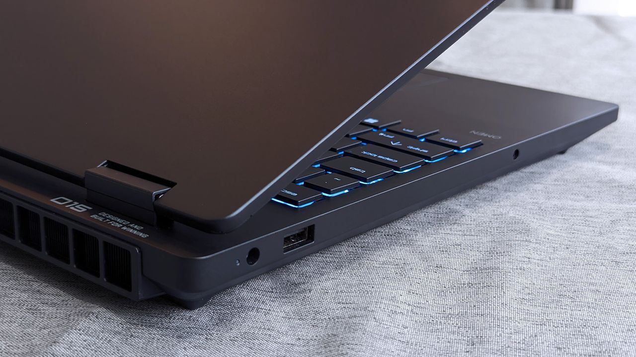 A partially open HyperX OMEN 15 laptop showing blue backlit arrow and modifier keys, along with visible side ports including USB and a headphone jack near the hinge.