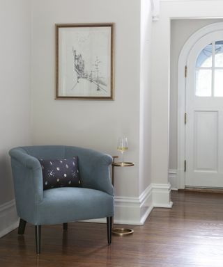 A foyer with white walls, a blue linen upholstered armchair and brass-framed artwork