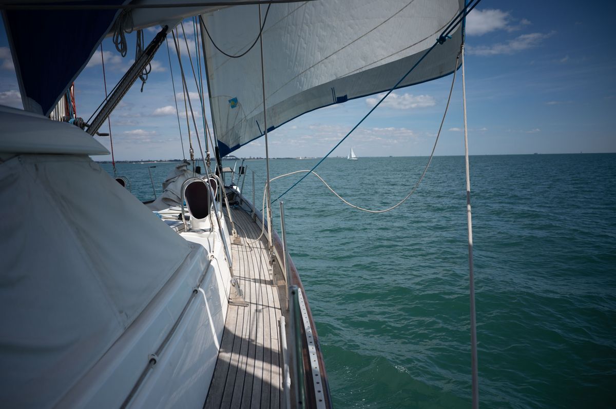 Side of a yacht from on board, there's a distant sail boat on the horizon, the water are calm, it's sunny