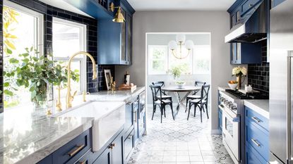 A small kitchen leading into a dining room with dark blue cabinets, marble countertops, brass taps and fixtures. 