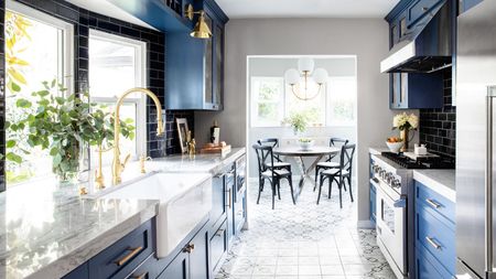 A small kitchen leading into a dining room with dark blue cabinets, marble countertops, brass taps and fixtures.