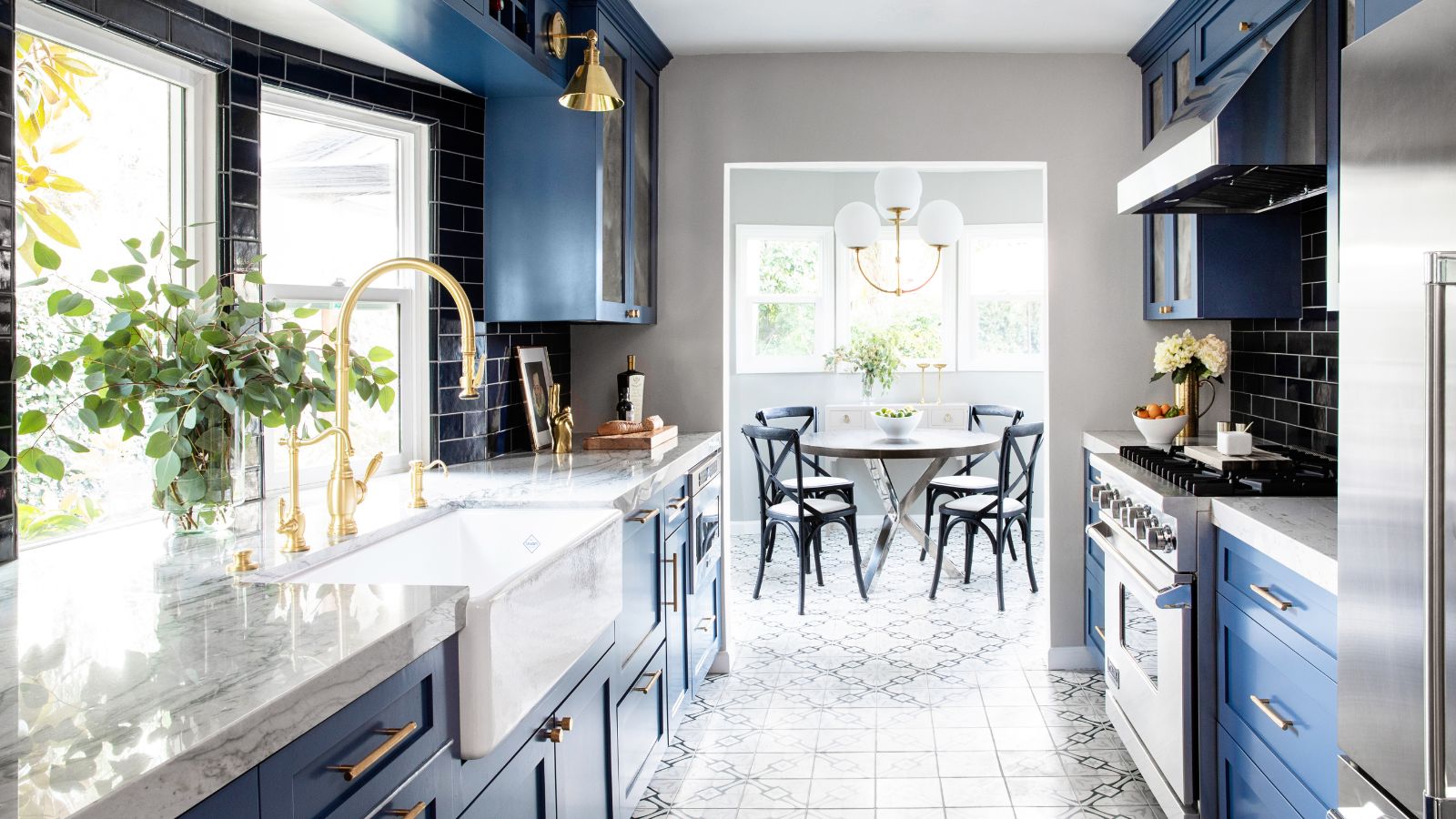 A small kitchen leading into a dining room with dark blue cabinets, marble countertops, brass taps and fixtures. 