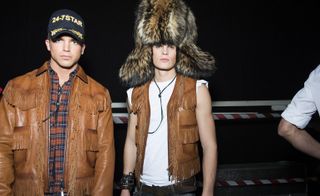 Two male models wearing looks from Dsquared2's collection. One model is wearing a brown fringed jacket, brown and blue plaid shirt and blue and gold hat with the wording '24-7STAR' embroidered on it. And the other model is wearing a white short sleeve top, brown fringed gilet and oversized fur hat