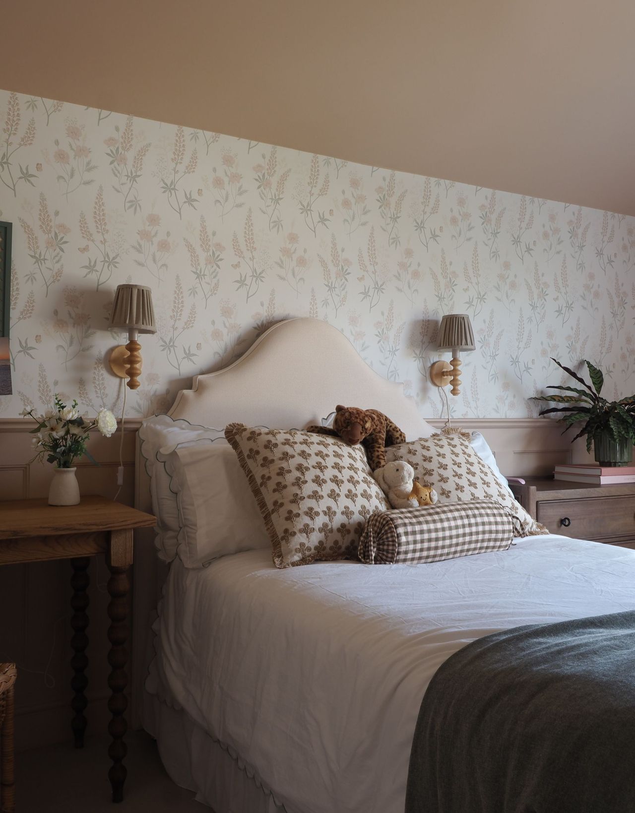 A modern bedroom with a peige-painted ceiling, wall lights wither side of a beige headbord and teddies and cushions on the bed