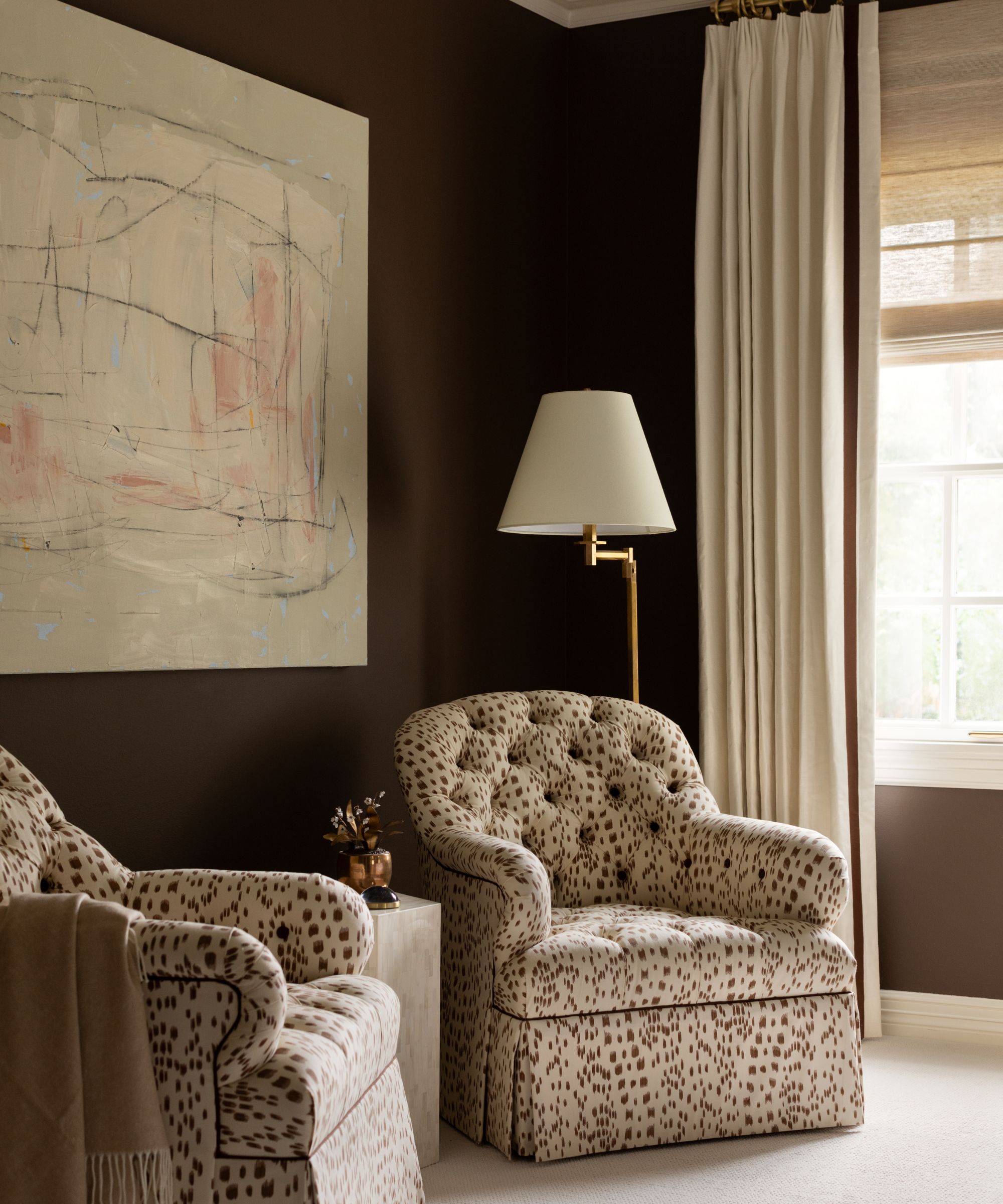 A bedroom with chocolate brown walls, a large piece of abstract artwork, cream curtains, and two neutral patterned armchairs.
