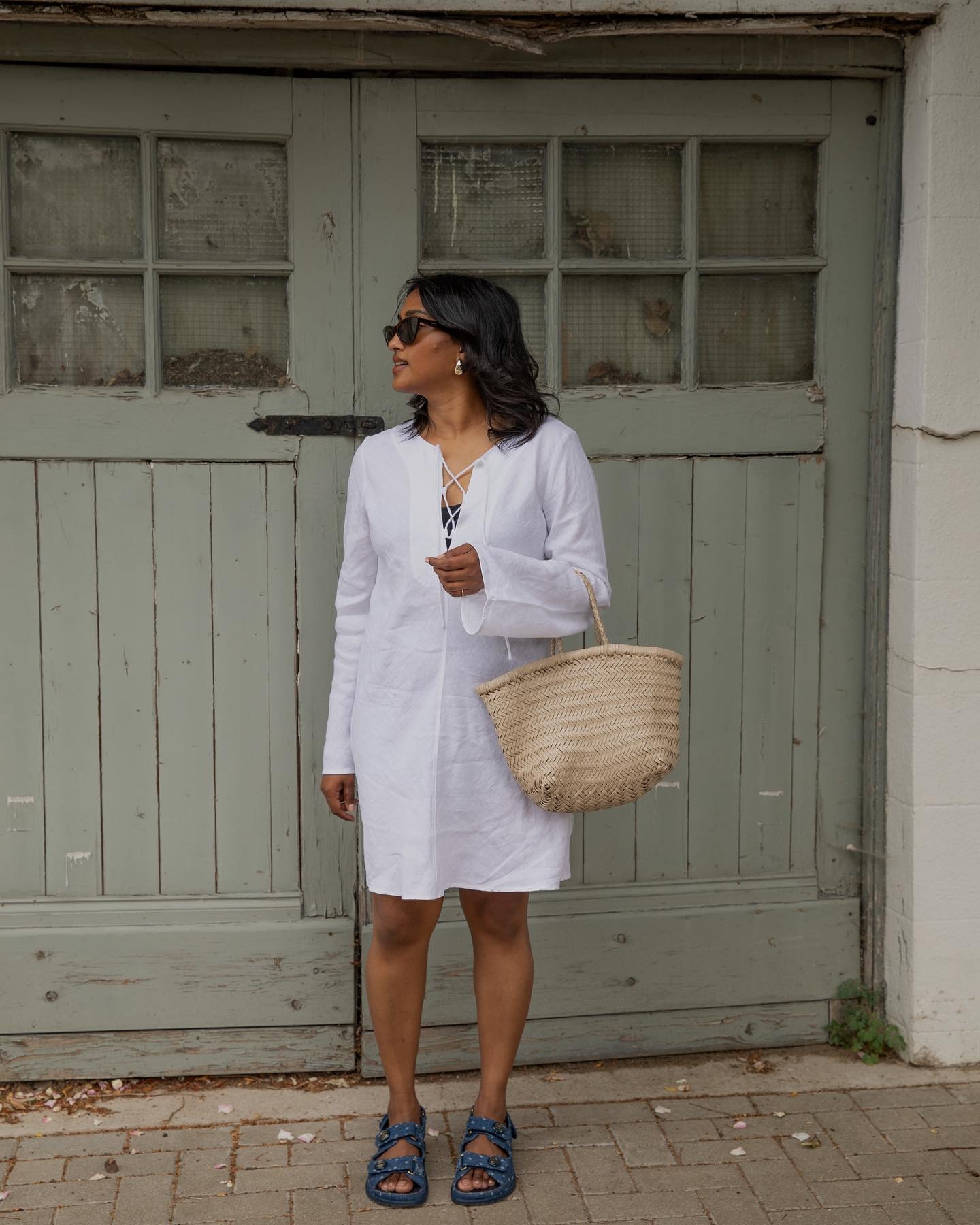 Woman wears white dress, denim sandals, holds cream leather woven bag