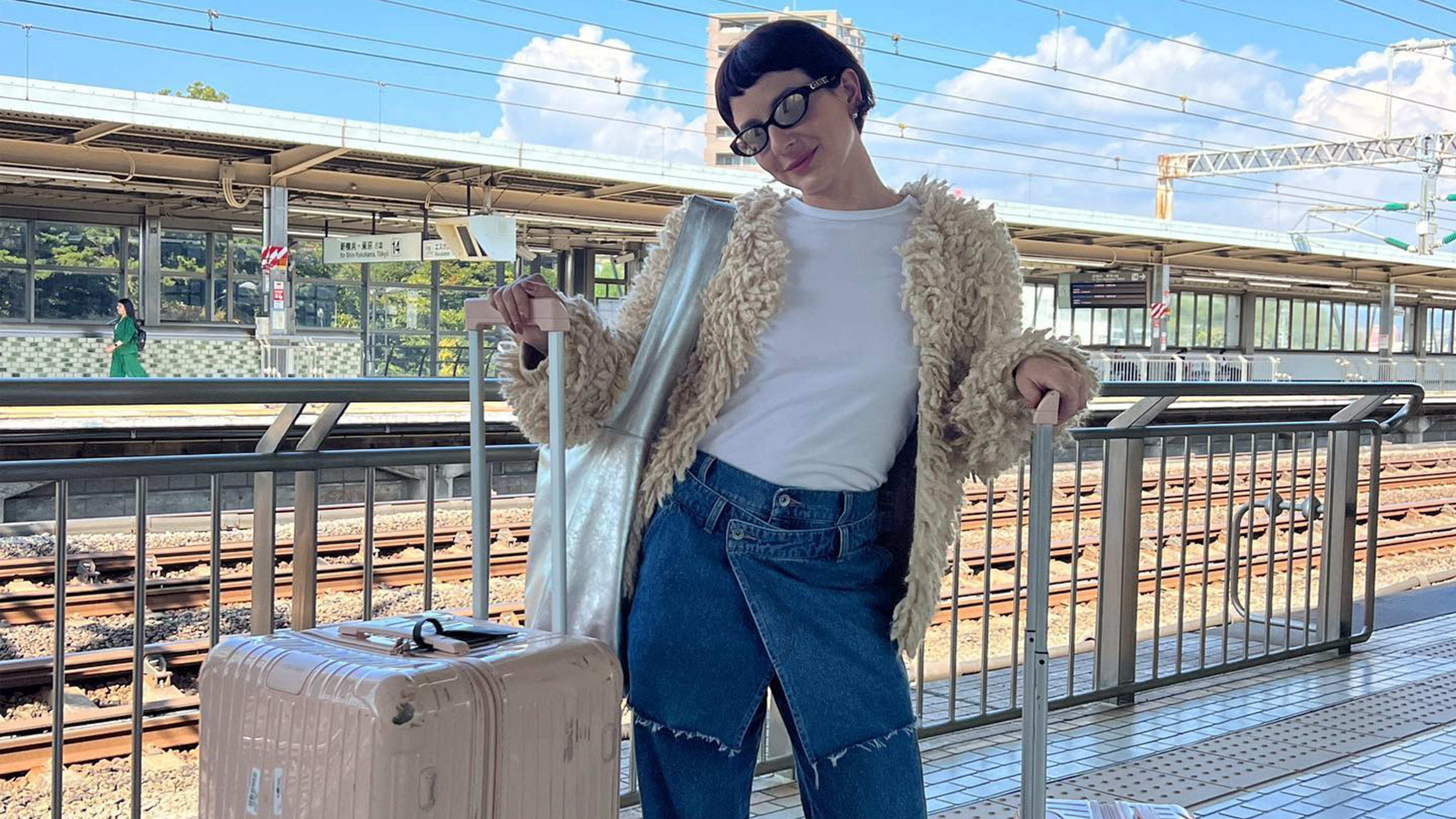 Alyssa wears a fur jacket, white tee, and jeans, with two suitcases.