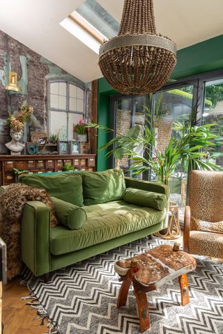 A living room with green wall paint decor, green suede sofa, palm plants and zig-zagged rug