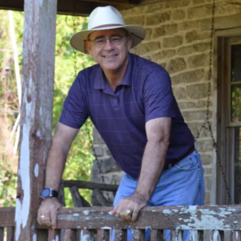a photo of gardening expert Skip Richter: a man standing on a wooden balcony next to a tree in blue shorts, a navy short sleeved shirt, thin framed metal glasses and a white trilby hat smiling at the camera