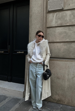 what to wear in 30-degree temperatures, shown in a photo of a woman standing outside wearing a white leather trench coat with a fur collar over a cashmere hoodie with baggy jeans and silver boots