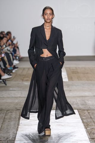 A female model wearing a black long sleeve shirt with a cape, black pants and platform shoes walking down a runway.