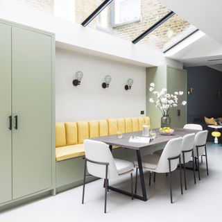 White dining room with sage green cabinetry, yellow upholstered in built bench by dining table