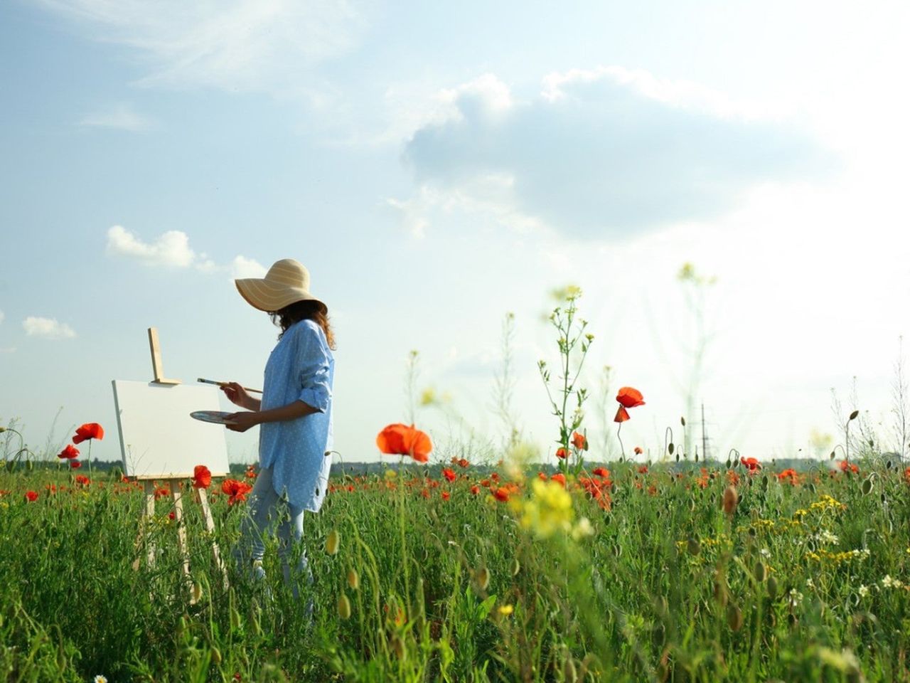 A woman in a sun hat paints at an easel in a field of red poppies