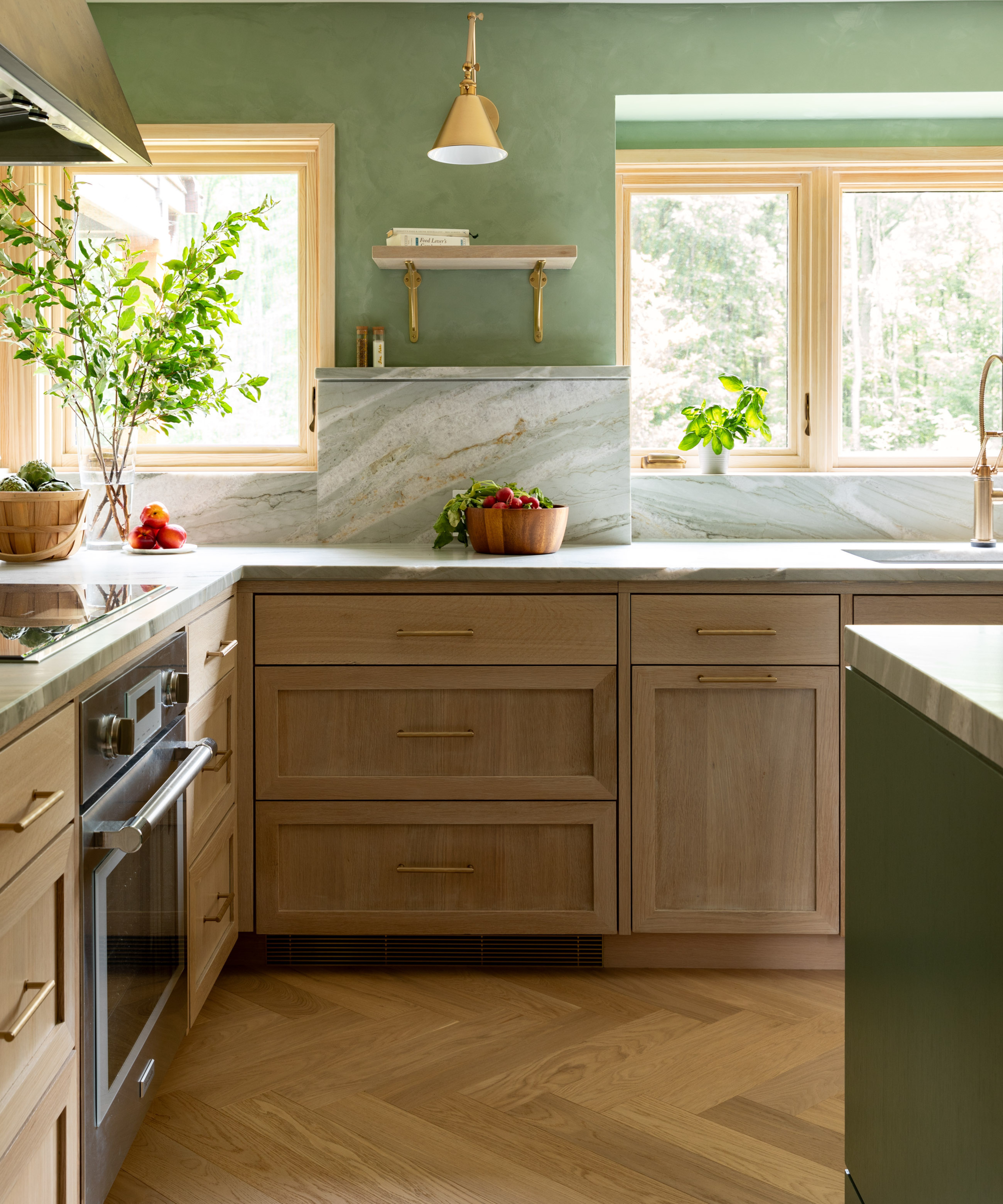 A kitchen with sage green plastered walls, light wood cabinetry, and a marble backsplash that steps up behind the counter, all set against a light oak herringbone floor
