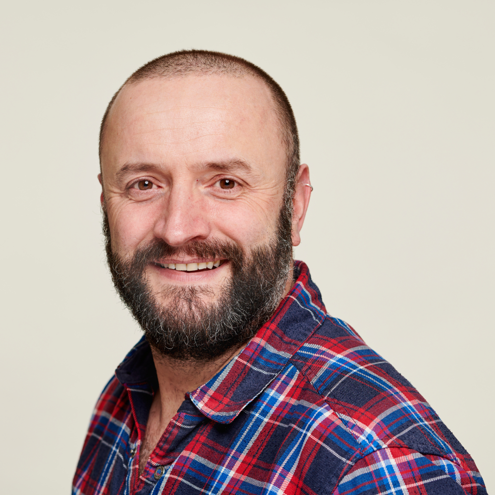 male with short dark hair and beard wearing blue, red and white checked shirt