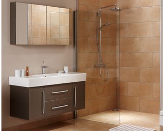 Warm sand colored tiles line the floor, shower and walls in this modern, sleek bathroom. There is a mounted mirrored wall cabinet on the left above a wide floating sink and drawers