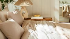 Beige Brooklinen bedding styled in a sunlit neutral bedroom that features a pleated table lamp and stack of books
