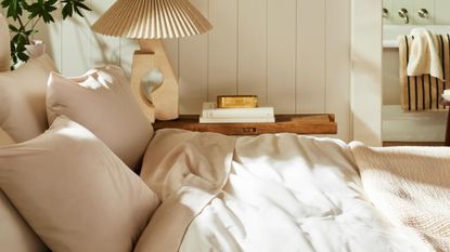 Beige Brooklinen bedding styled in a sunlit neutral bedroom that features a pleated table lamp and stack of books