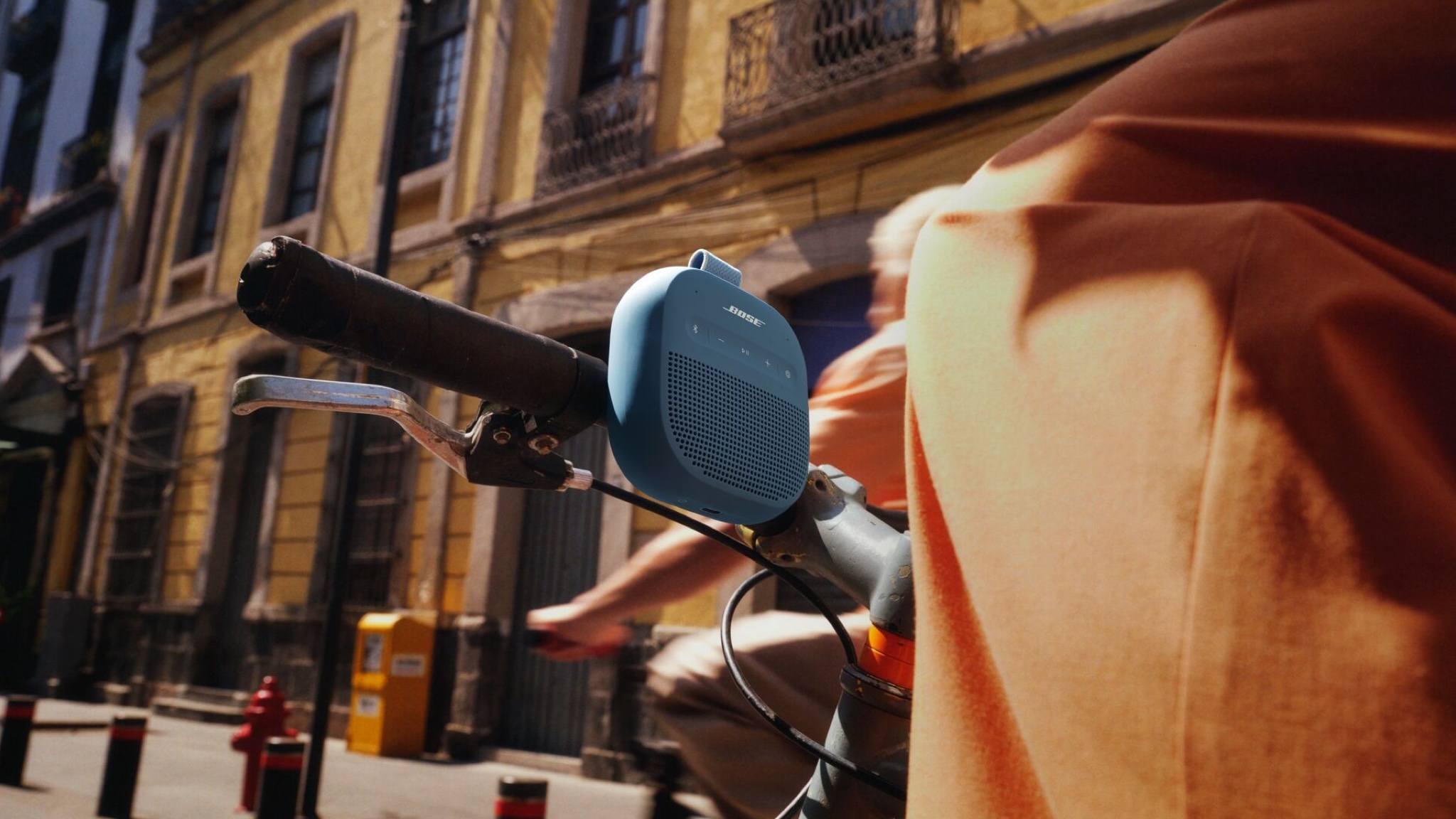 A blue SoundLink Micro speaker attached to the handlebars of a bicycle in a street of what look like sandstone flats or townhouses. The bicycle is moving but we can't see the rider.