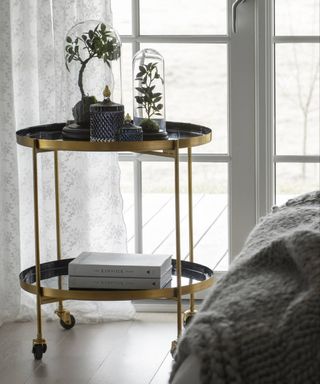 A brass bar cart in the bedroom with smoked grey glass jar, white hardback books and plant