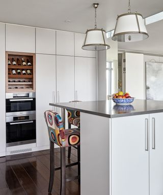 A kitchen with white cabinets, a white island with a metallic worktop and multi colored bar stools