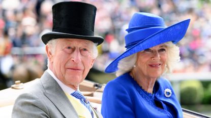 King Charles wears a gray suit with a black top hat, while Queen Camilla wears a large royal blue hat with a matching jacket