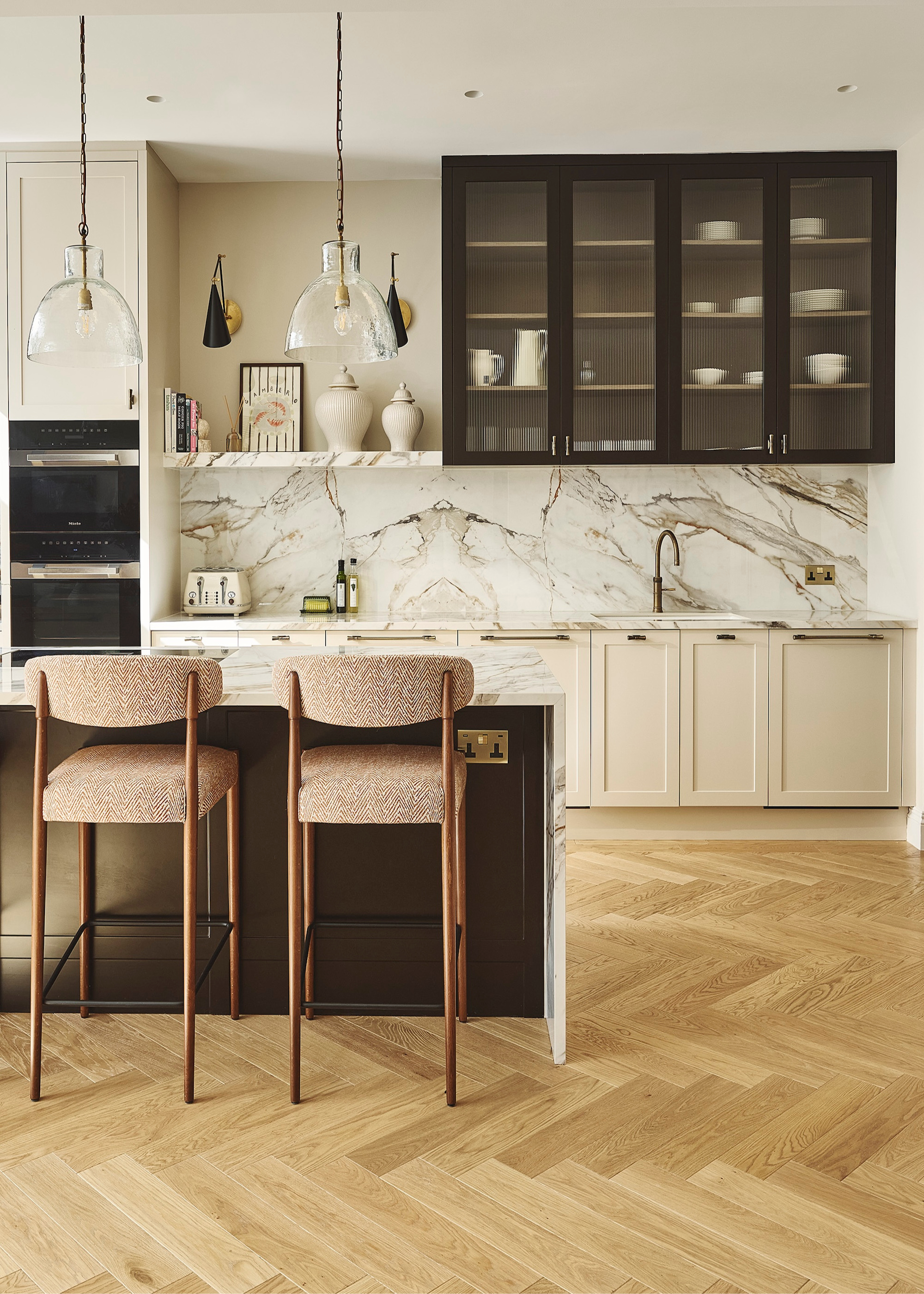 A neutral kitchen with herringbone wood flooring, black-framed and glass-fronted kitchen cabinets above with cream lower cabinets, and glass pendant lights hang above the kitchen island of the same veined stone used as the kitchen's backsplash