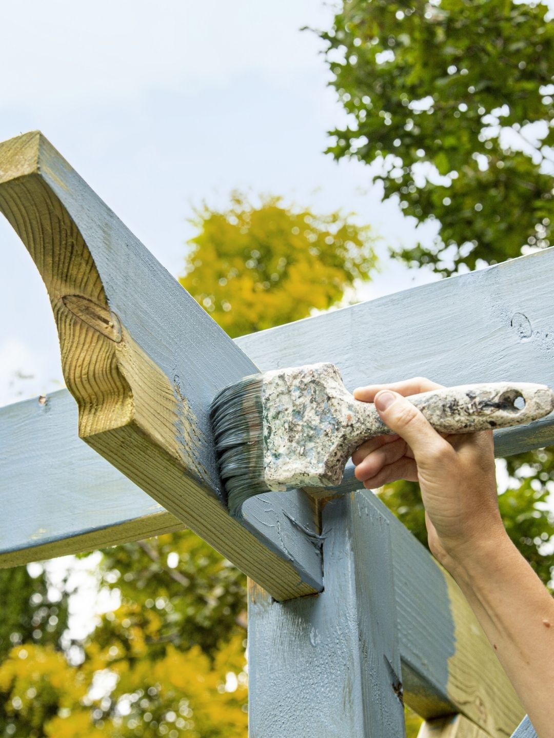 Image of a hand holding a paint brush and painting an outdoor pergola a light blue/gray color.