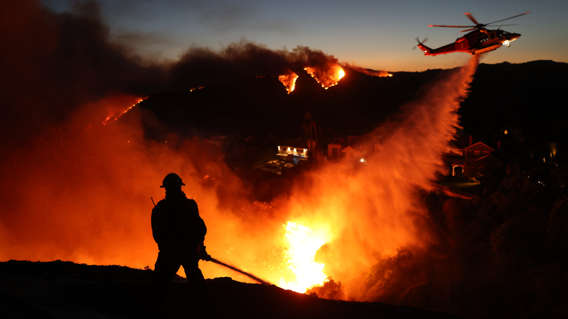A firefighter is silhouetted in the glowing orange and yellow blaze of a wildfire as a helicopter above dumps a stream of water below