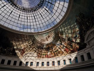 The Bourse de Commerce in Paris. Large, circular hall, with renaissance art on the walls and a glass dome. Light coming through the dome casts a shadow on the walls.