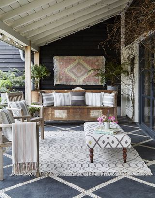 front porch with vintage bench, wood armchairs, upholstered ottoman, pillows, blankets, rug, slate and pebble tiled floor, rug on wall, plants