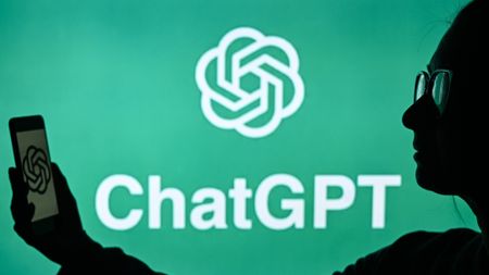 A woman holds a cell phone in front of a computer screen displaying the ChatGPT logo.