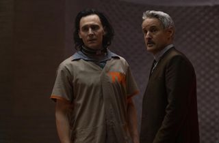 A captured and collared Loki (Tom Hiddleston) wears a beige TVA shirt while receiving instructions from besuited Mobius M Mobius (Owen Wilson)