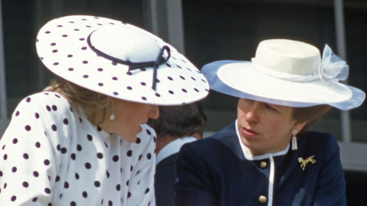 Princess Diana wearing a white polka dot dress and matching hat leaning forward to talk to Princess Anne wearing a blue jacket and white wide brimmed hat