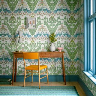 home office with bold blue and green patterned wallpaper, vintage desk and chair, blue painted woodwork, rug, artwork, lamp, jug and bowl