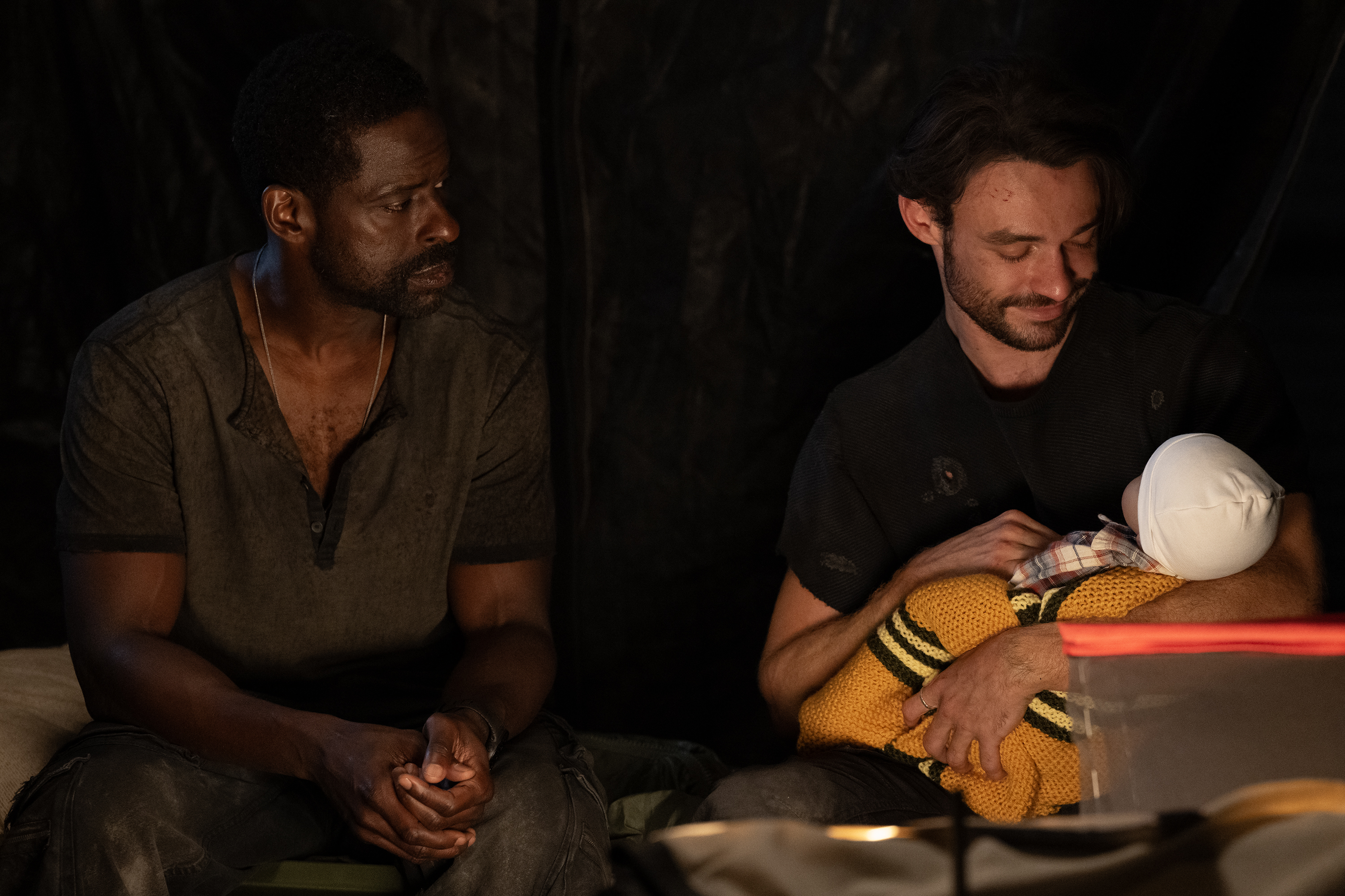 A black man (Sterling K. Brown as Xavier Collins) sits with a white man (Thomas Doherty as Link) who holds and gazes at a baby swaddled in a yellow blanket with stripes. Both the men sit outdoors at night, in a still from the 'Paradise' season 2 finale.