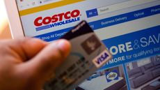 The Costco Wholesale Corporation website is displayed on a laptop in the background with a hand holding a bank card.