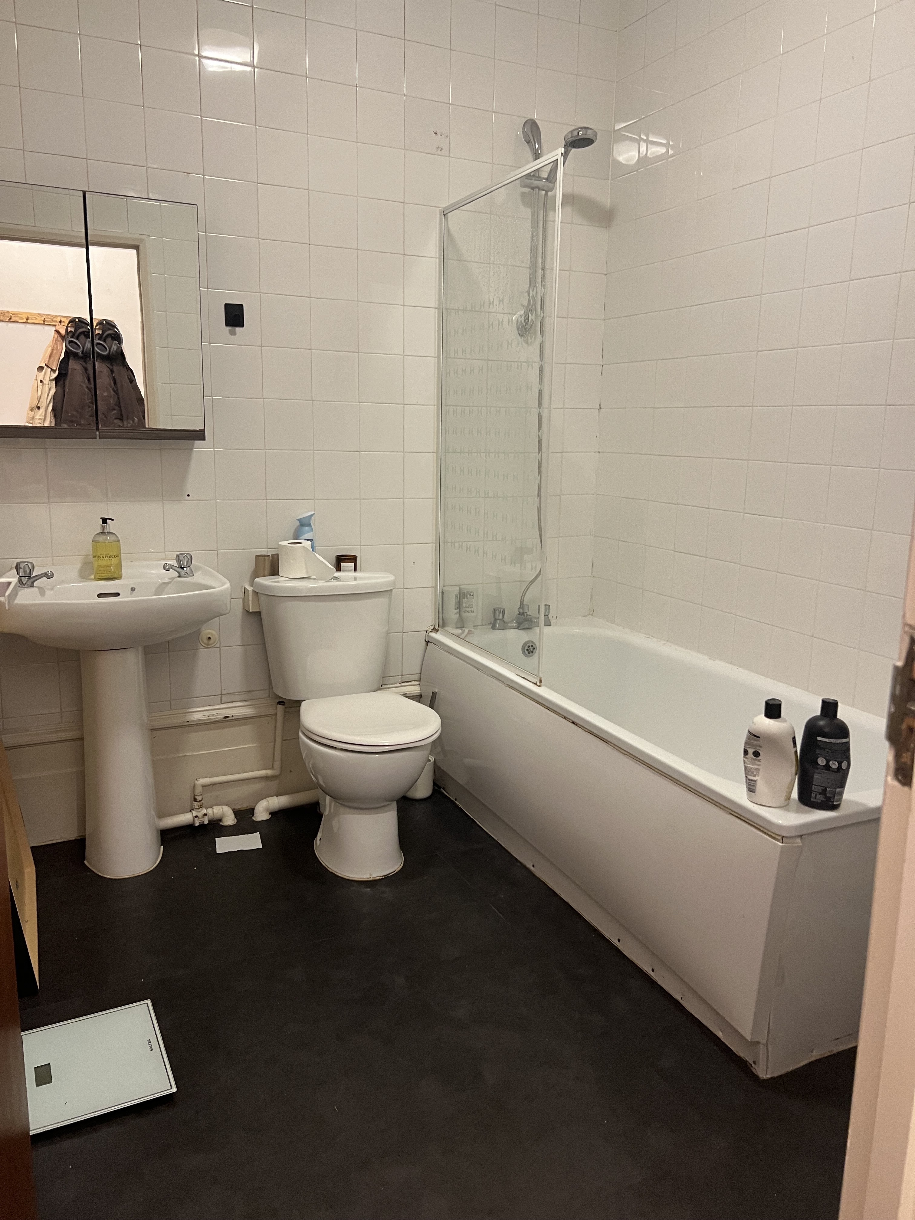 Image of a bathroom with black flooring and white, tiled walls. There is a white tub/shower, a white toilet, and a white sink.