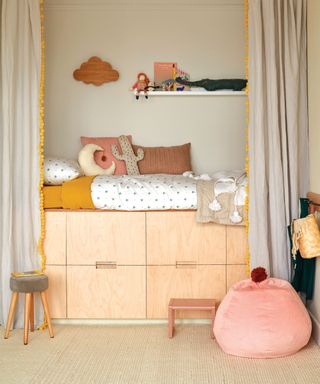 painted kids bedroom in beige-gray, matching beige-gray curtains, light wooden bedframe and storage, ochre and pink accents on cushions, bean-bag and accessories, wall mounted white shelf