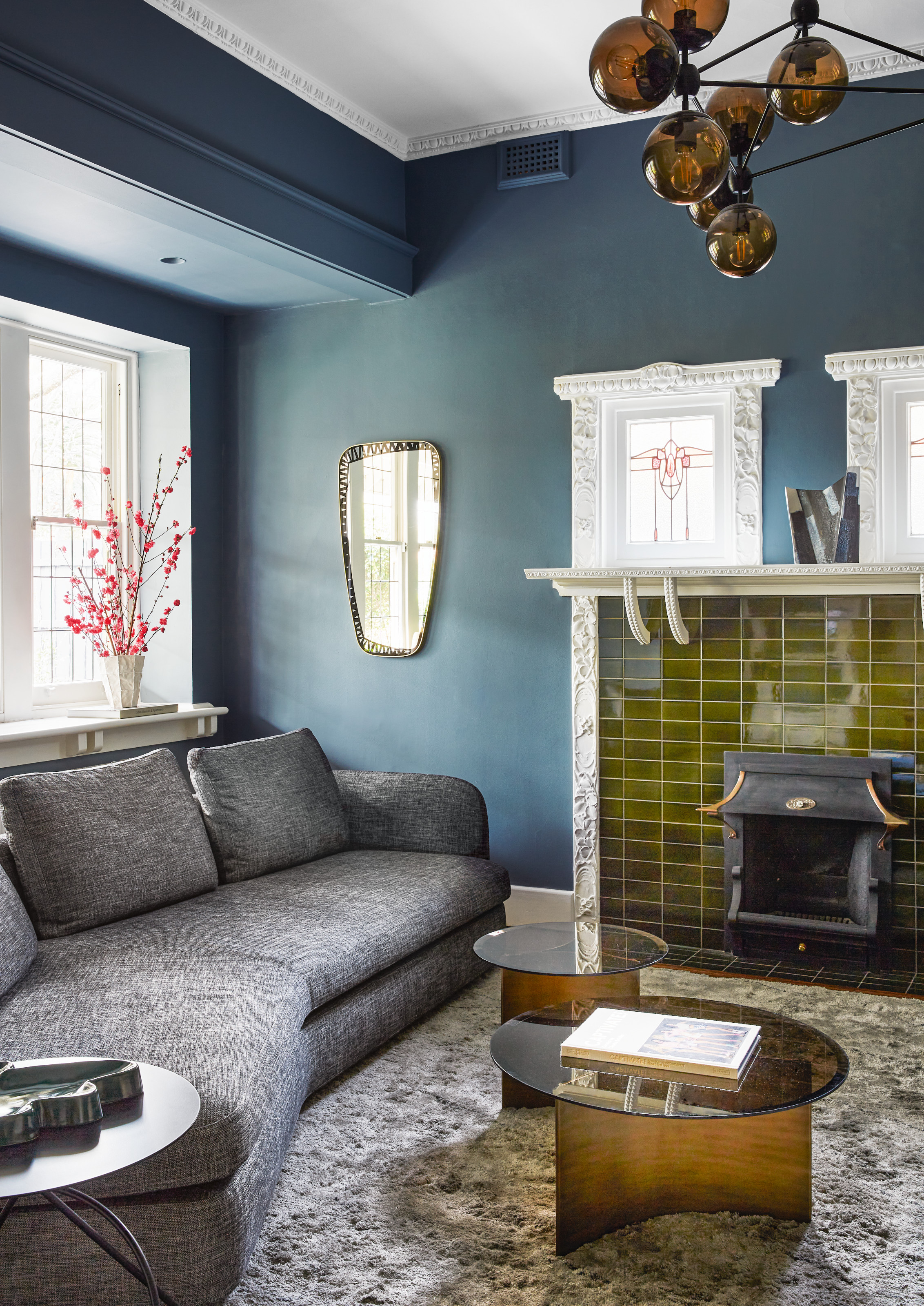 Living room with dark blue walls, grey angled sofa, fireplace with olive green tiles, and amber glass-topped round coffee tables