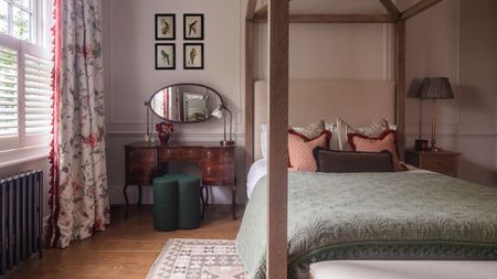 A traditional bedroom with a wooden dressing table and green stool beside a four poster bed. Wooden flooring and pink walls. 