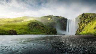 Skogafoss Waterfall in Iceland. Surrounded by green landscape and leads into a river.